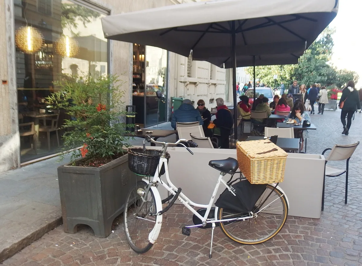 Weißes Stadtfahrrad mit Weidenkorb vor einer Caféterrasse in Parma geparkt