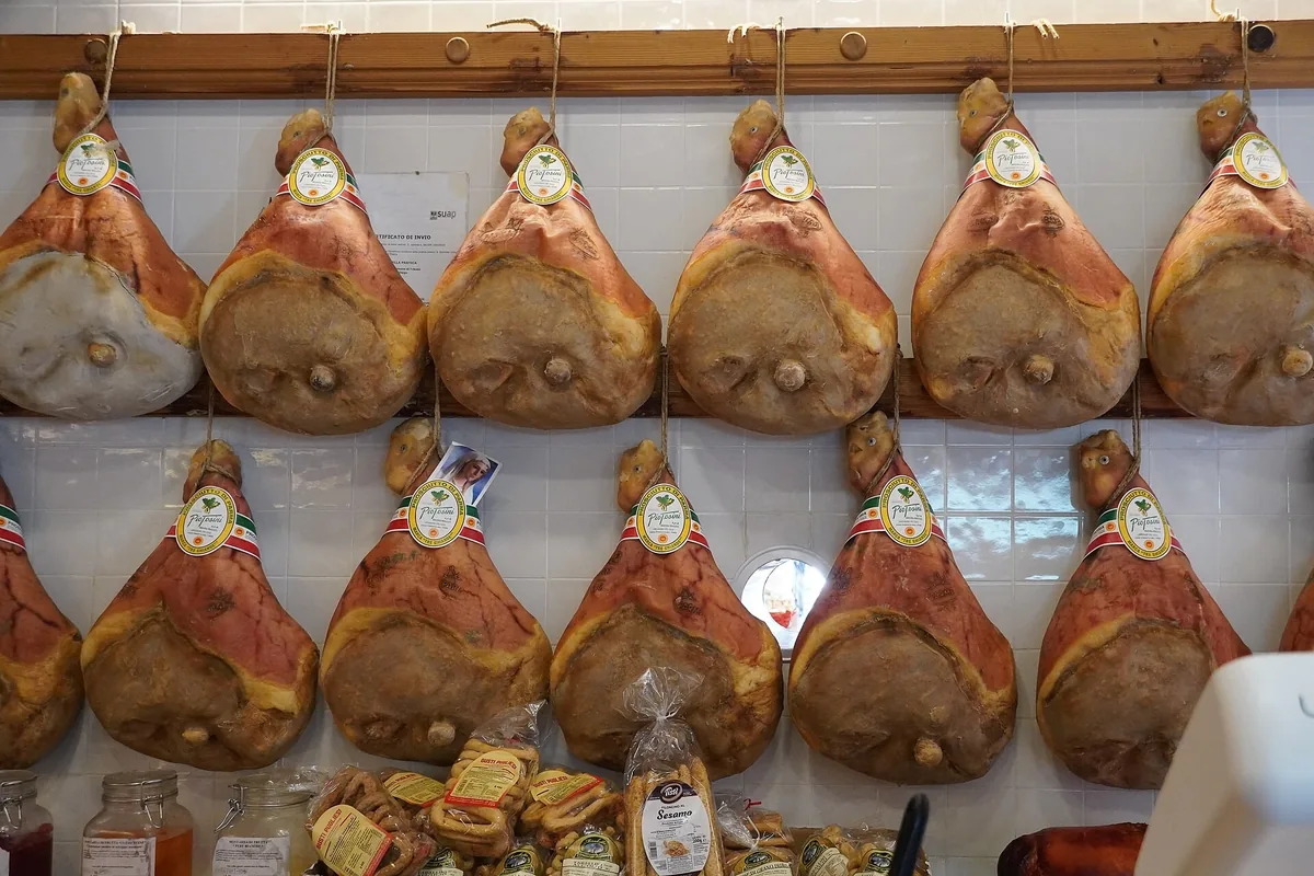 Zwei Reihen ganzer Prosciutto-di-Parma-Schinken, die an einer weißen Fliesenwand hängen